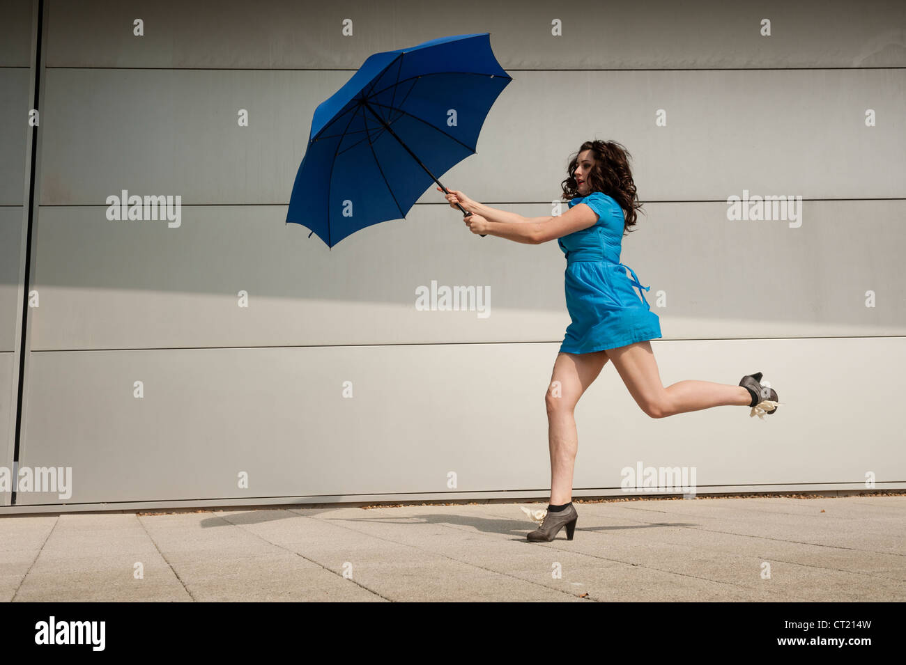 Une jeune femme seule fille portant une robe bleue courte fonctionnant sous un parapluie bleu parasol, UK Banque D'Images