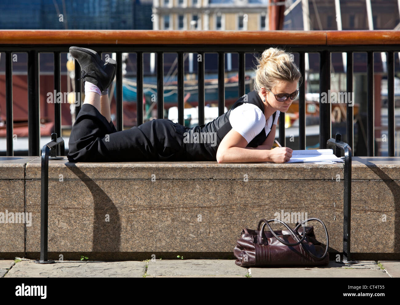 Portrait complet d'une femme couché sur un banc en position ventrale, écrivant sur un carnet, South Bank, Londres, Angleterre, Royaume-Uni. Banque D'Images