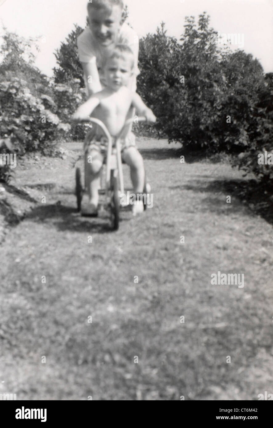 Photo en noir et blanc un jeune garçon poussant un jeune garçon sur son tricycle à l'extérieur dans le jardin album de famille Banque D'Images