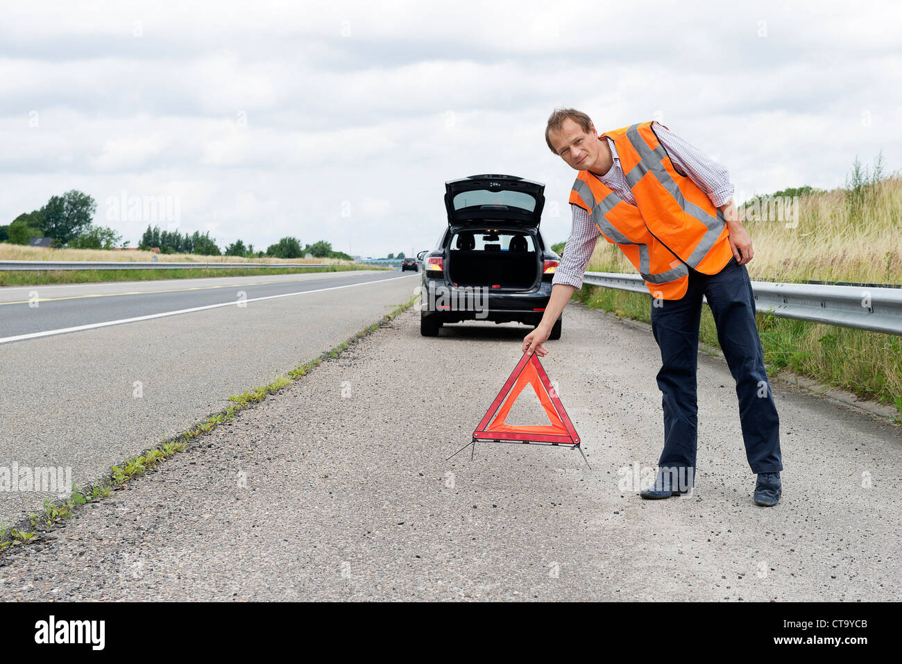 Automobiliste, portant un triangle d'avertissement, le plaçant à une distance sûre de sa voiture en panne le long d'une autoroute Banque D'Images