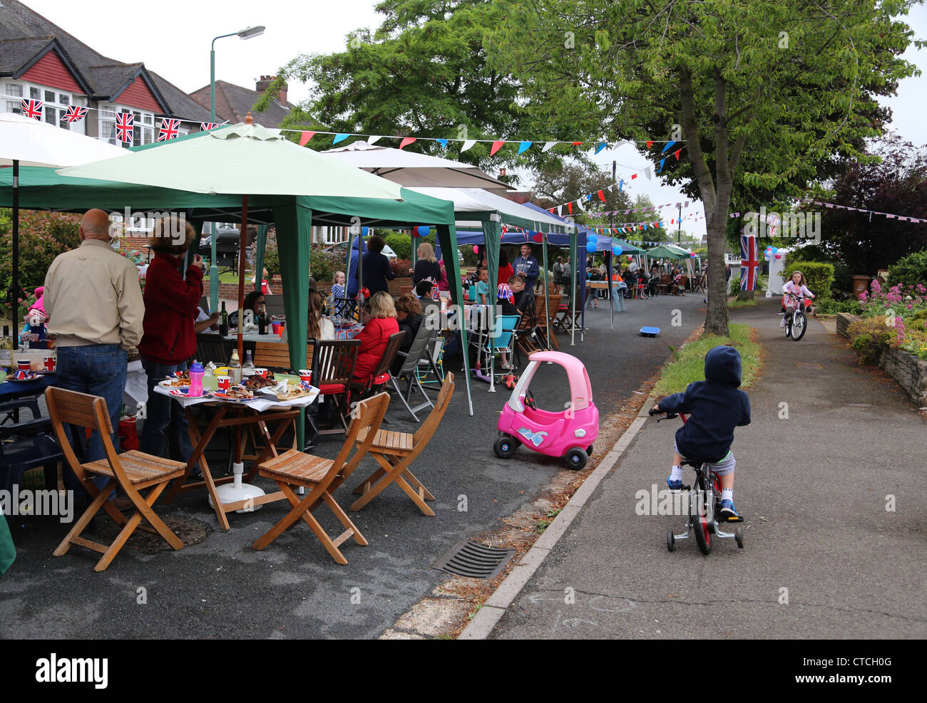 Les gens assis à l'ombre des parasols et chapiteaux de manger et de boire pendant que les enfants sont la bicyclette à Street Party Banque D'Images
