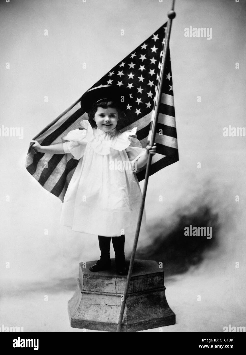 1890 TOUR DE SIÈCLE SMILING LITTLE GIRL STANDING ON PLATFORM VÊTUE D'UNE ROBE BLANCHE ET UN CHAPEAU SOMBRE tenant un drapeau américain Banque D'Images