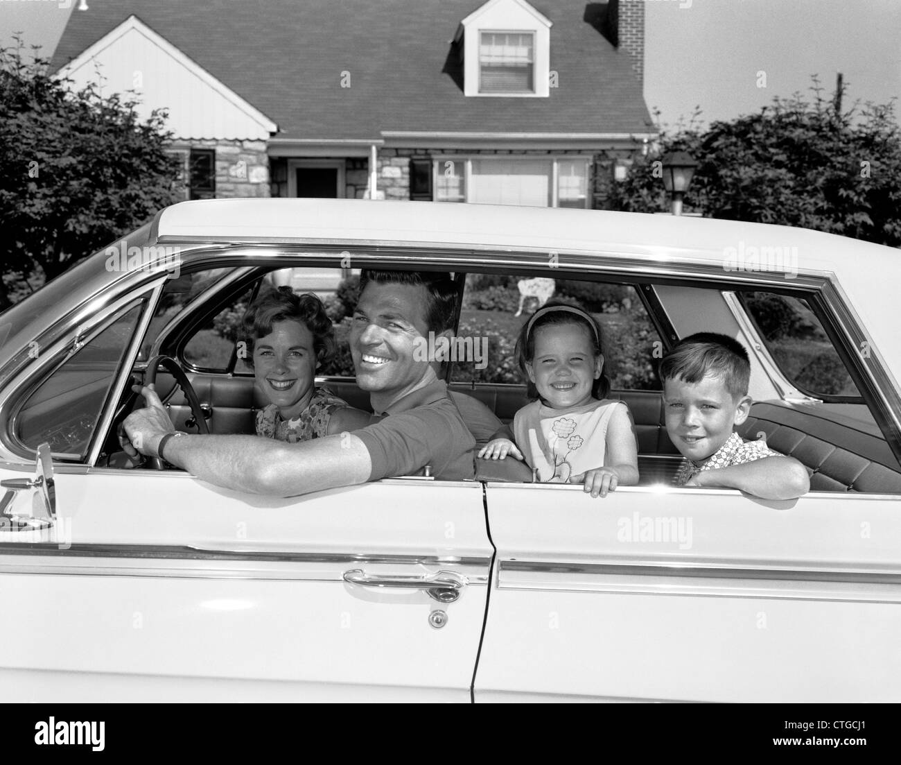 1960 SMILING FAMILY Père Mère Fils Fille ASSIS DANS LA VOITURE EN FACE DE SUBURBAN HOUSE Vue de côté à l'extérieur Banque D'Images