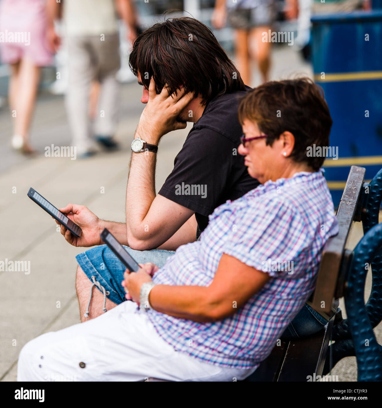 Un homme femme couple lecture des e-books sur leurs lecteurs Amazon Kindle, assis dehors sur une journée chaude, UK Banque D'Images