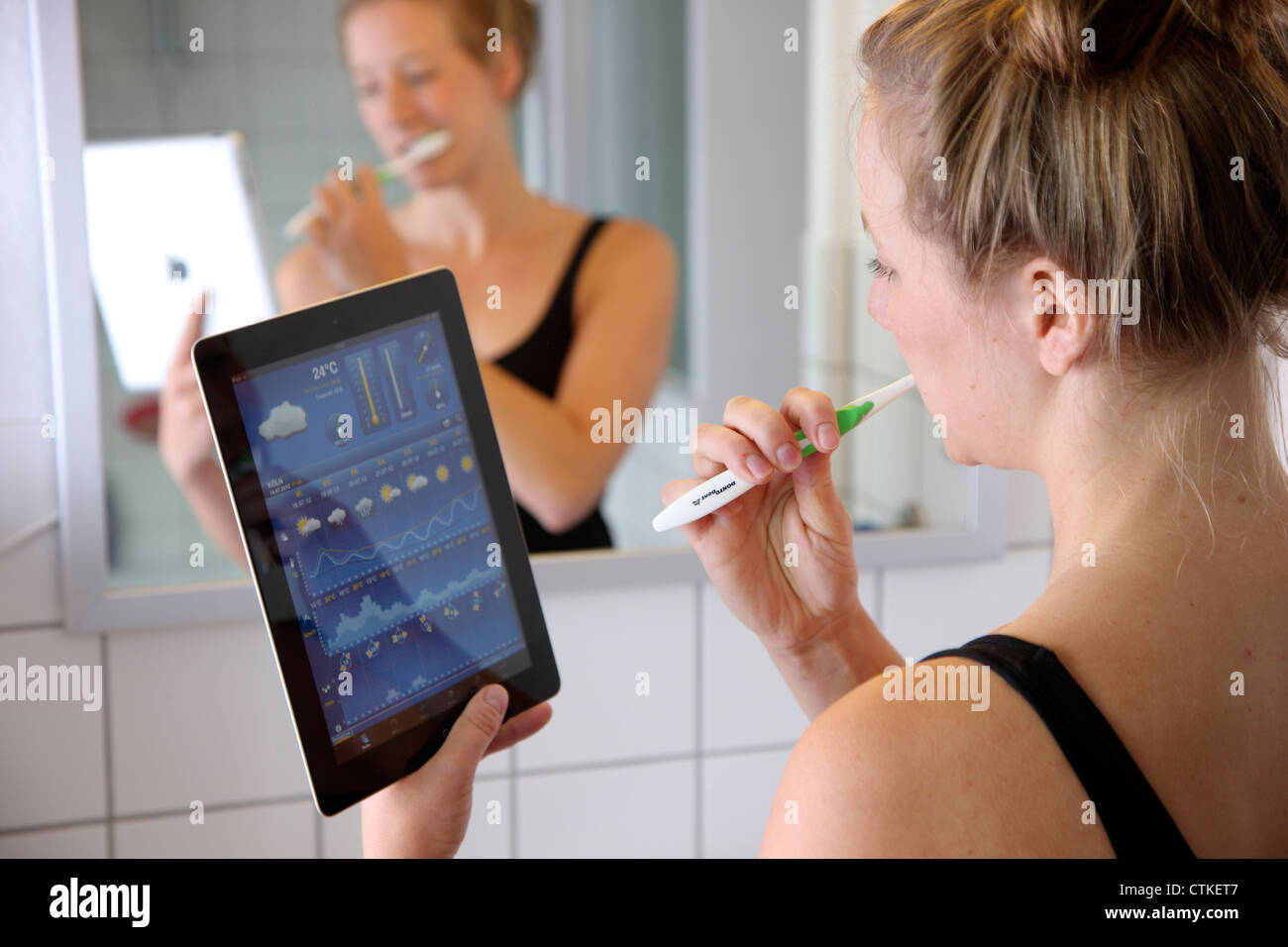 Jeune femme dans la salle de bain, se brosser les dents, à l'aide d'une tablette IPad, ordinateur, lecture, prévisions météo. Banque D'Images