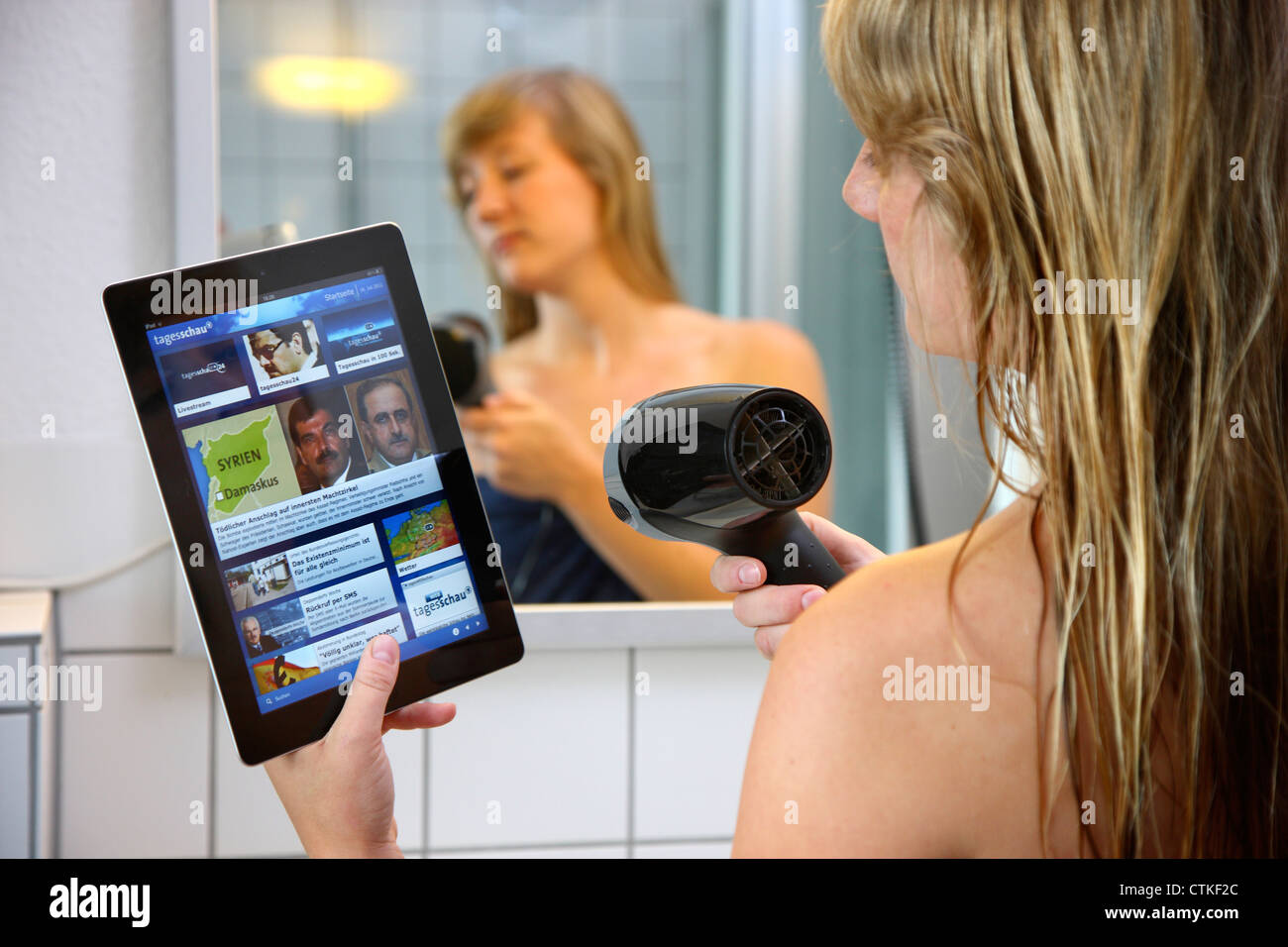 Femme est en train de sécher ses cheveux dans la salle de bains, à l'aide d'un ordinateur tablette, l'Ipad, la lecture de l'actualité sur l'Internet. Banque D'Images