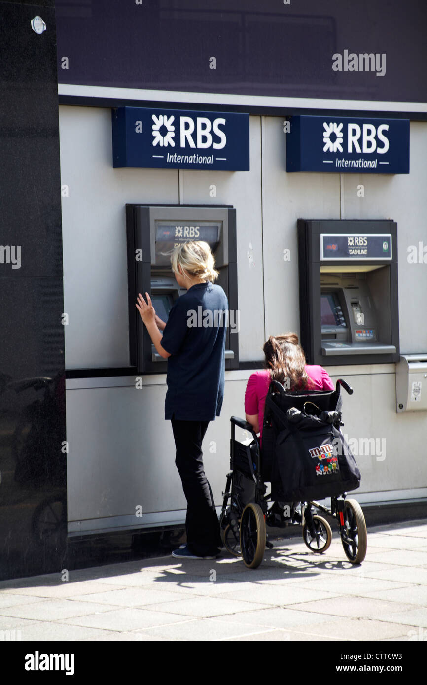 Jeune femme à l'aide de RBS International cash machine tandis qu'un autre attend aux côtés de fauteuil roulant à St Helier, Jersey en Juillet Banque D'Images