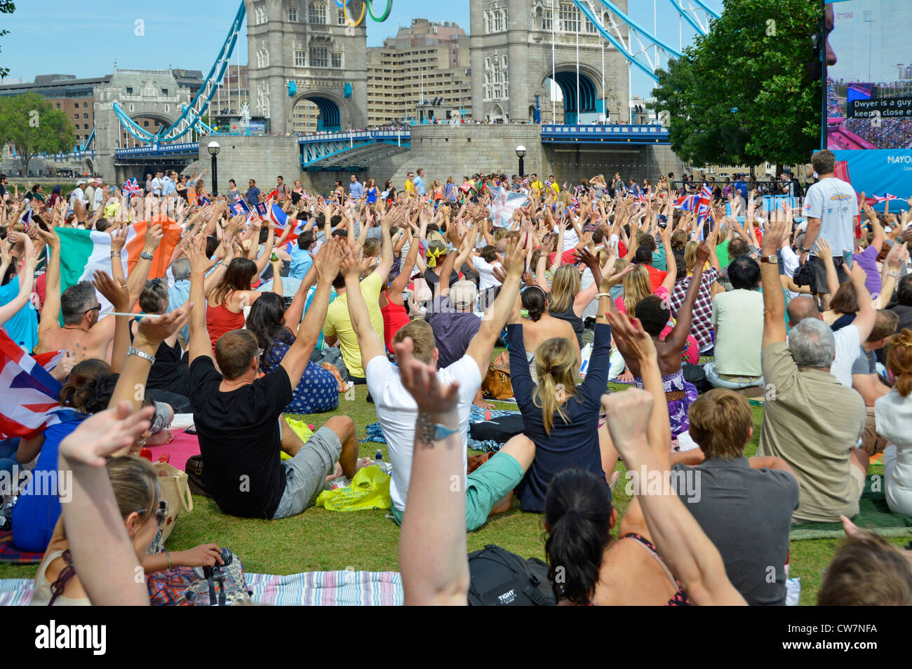 Les gens de foule s'assoient sur la pelouse des Jeux olympiques de Londres 2012 Téléviseur grand écran montre les mains vers le haut et le pont de la tour Potters Fields Park Southwark Angleterre Royaume-Uni Banque D'Images