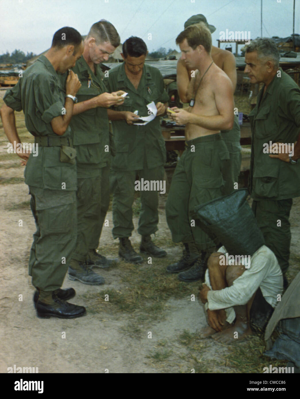 Guerre du Vietnam. On soupçonne un Viet Cong s'assied sur le sol avec un sac noir sur la tête, tandis que plusieurs soldats américains d'examiner des documents. Banque D'Images