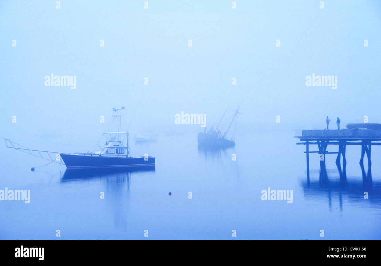 Deux hommes entourent la pêche par le brouillard et debout sur une jetée au petit matin avec un naufrage bateau et un bateau flottant à proximité. Banque D'Images