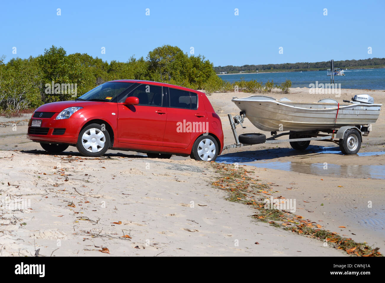 La Suzuki Swift rouge remorque bateau dans l'eau en face de la plage Banque D'Images