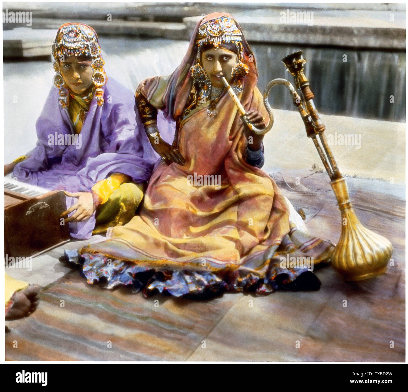 Portrait colorisé de deux femmes, vêtues d'une tenue opulente, assis sur des tapis pendant un festival, Delhi, Inde, 1912. L'une des femmes fume un tuyau d'eau, tandis que l'autre joue un harmonium. (Photo de Burton Holmes) Banque D'Images