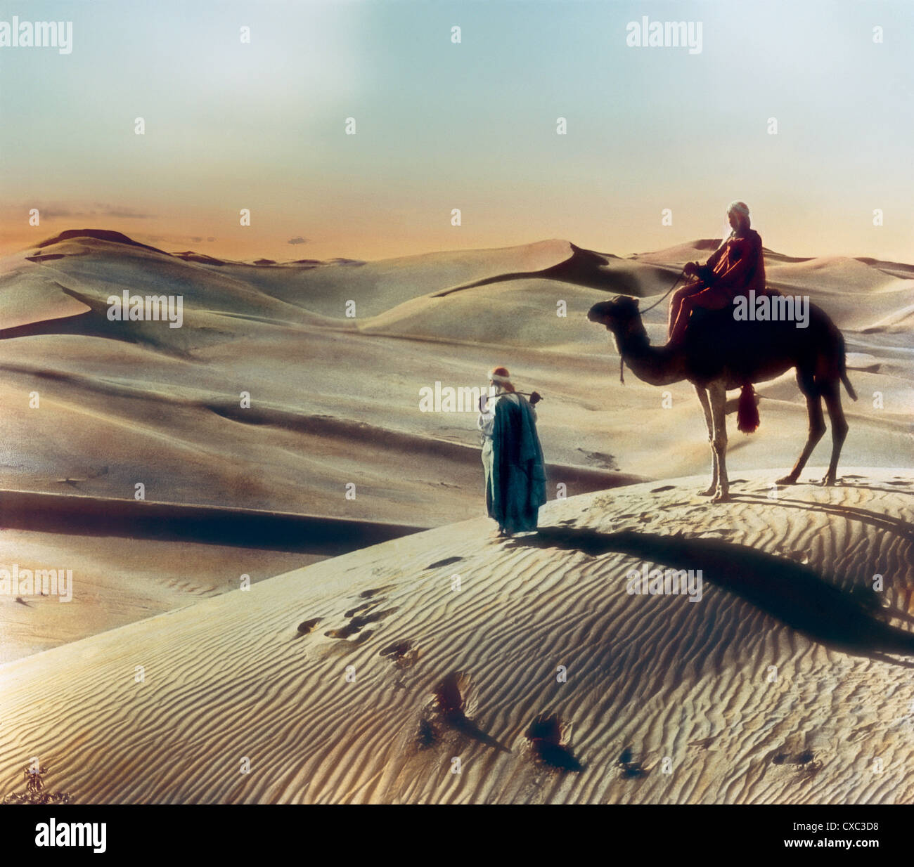 Photo colorisée d'une personne à cheval sur un chameau arrêtée sur une dune de sable dans le désert au coucher du soleil, Giza, Egypte, 1906. Un homme, peut-être un guide, se tient devant le chameau et tient un fouet sur son dos. (Photo de Burton Holmes) Banque D'Images