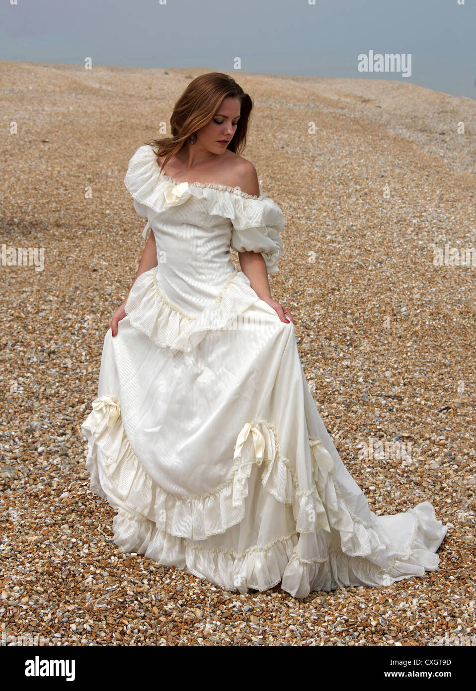 Jeune fille portant une robe de mariage blanc crème sur une plage de galets au bord de la mer. Banque D'Images