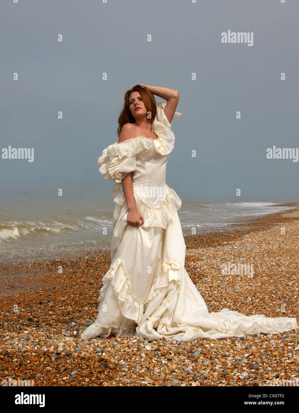 Jeune fille portant une robe de mariage blanc crème sur une plage de galets au bord de la mer. Banque D'Images