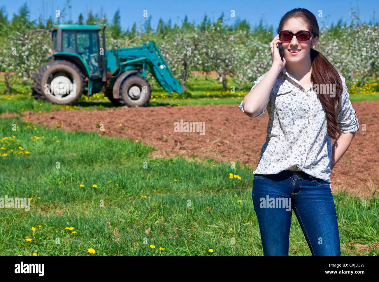 Jeune femme parlant l'agriculteur sur son téléphone avec un tracteur et un verger dans le champ derrière elle. Banque D'Images