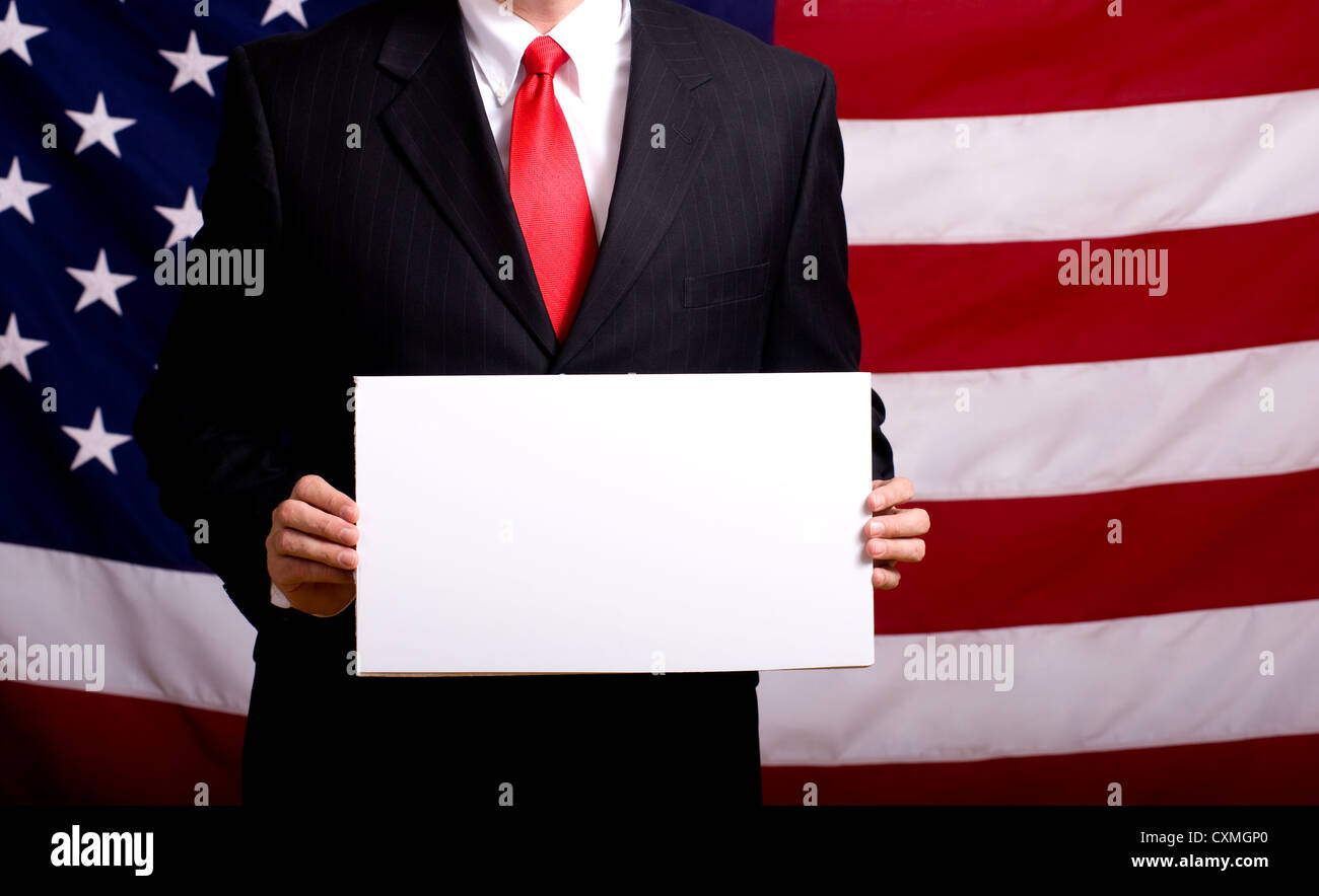 Un politicien ou homme d'affaires en costume holding a blank white signe devant le drapeau américain Banque D'Images