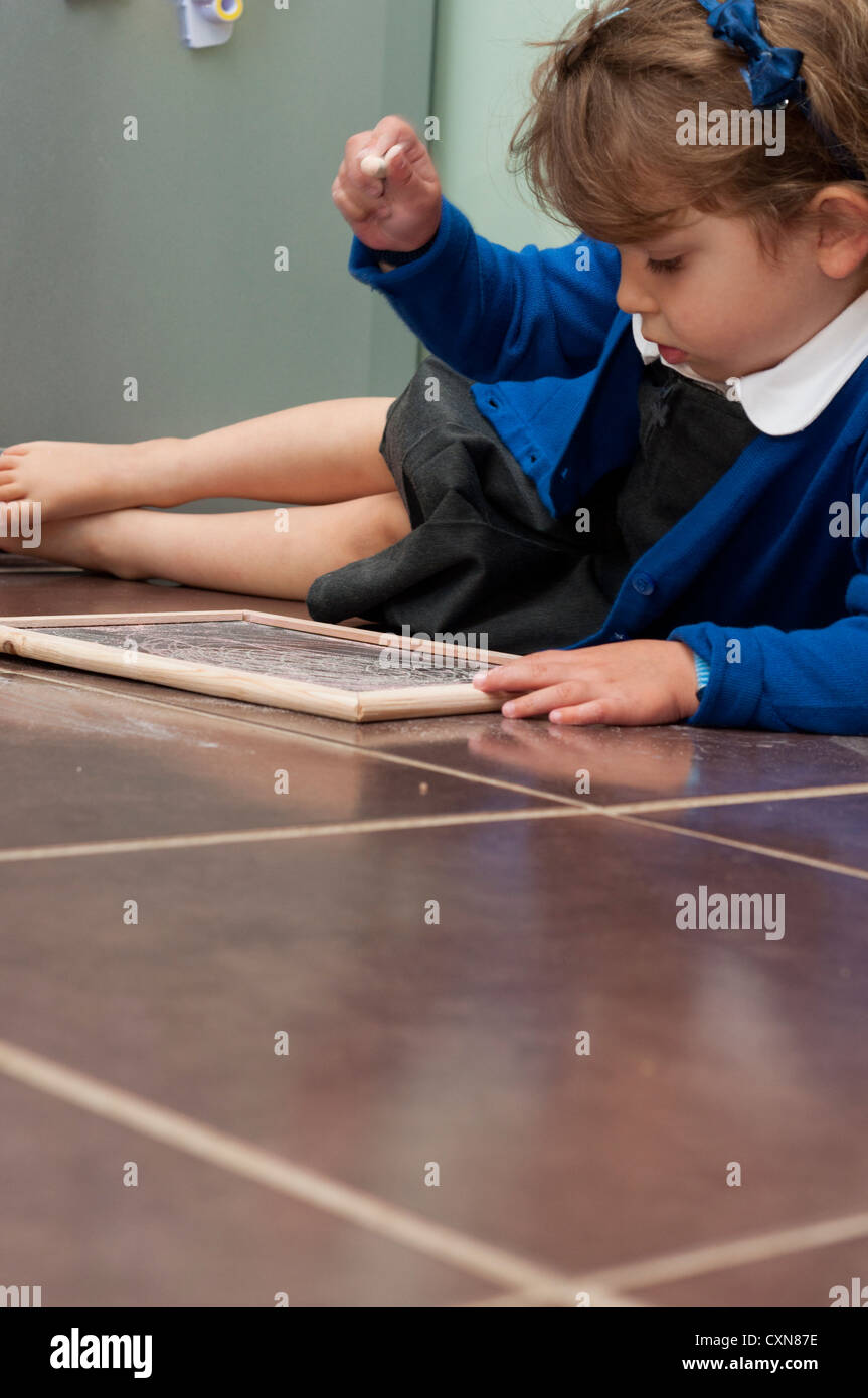 Quatre ans, fille en uniforme d'assis sur le plancher de la cuisine en s'appuyant sur un petit tableau. Banque D'Images