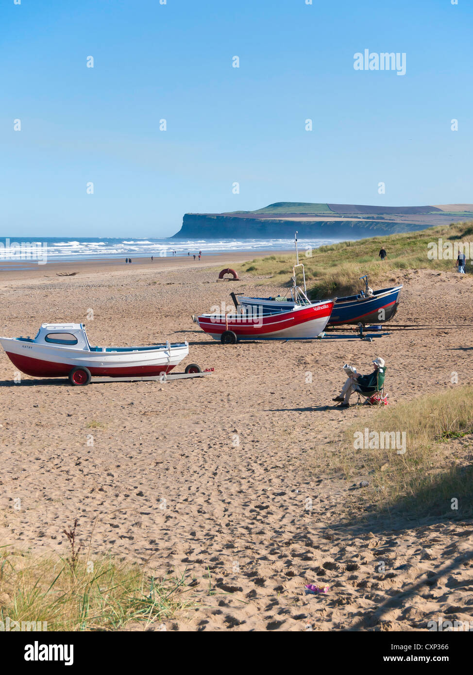 Sous le soleil d'automne dimanche après-midi, un homme lit un journal par les bateaux de pêche sur la plage de Marske Banque D'Images
