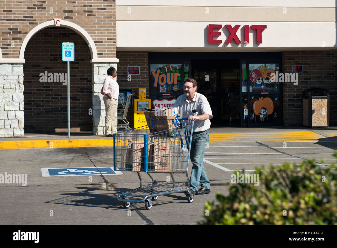 Un homme de race blanche dans son 50s pousse un panier avec sac la nourriture dans un parking en face d'un supermarché. Banque D'Images