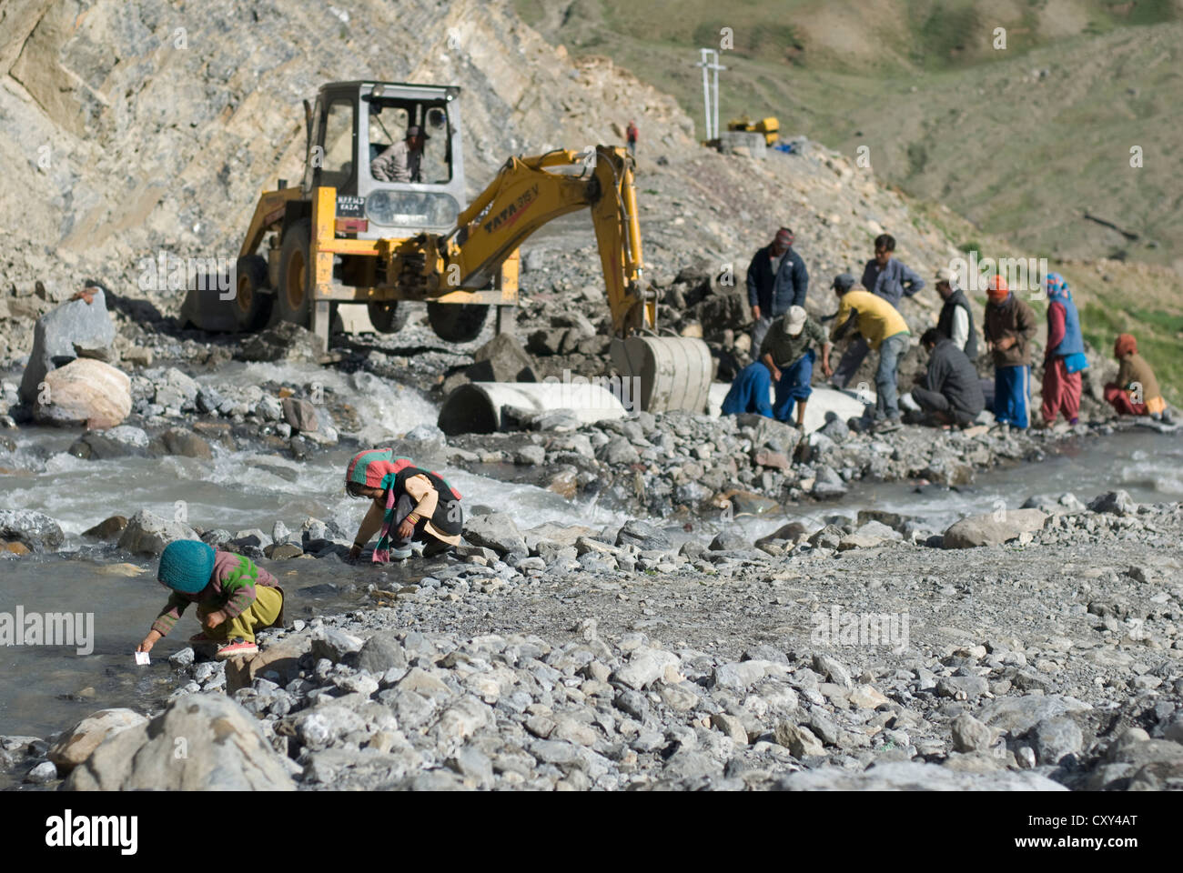 Les enfants jouent au bord de la rivière comme une masse-mover travaille pour effacer une route endommagée par les inondations dans le village de boue, le Spiti, le nord de l'Inde Banque D'Images