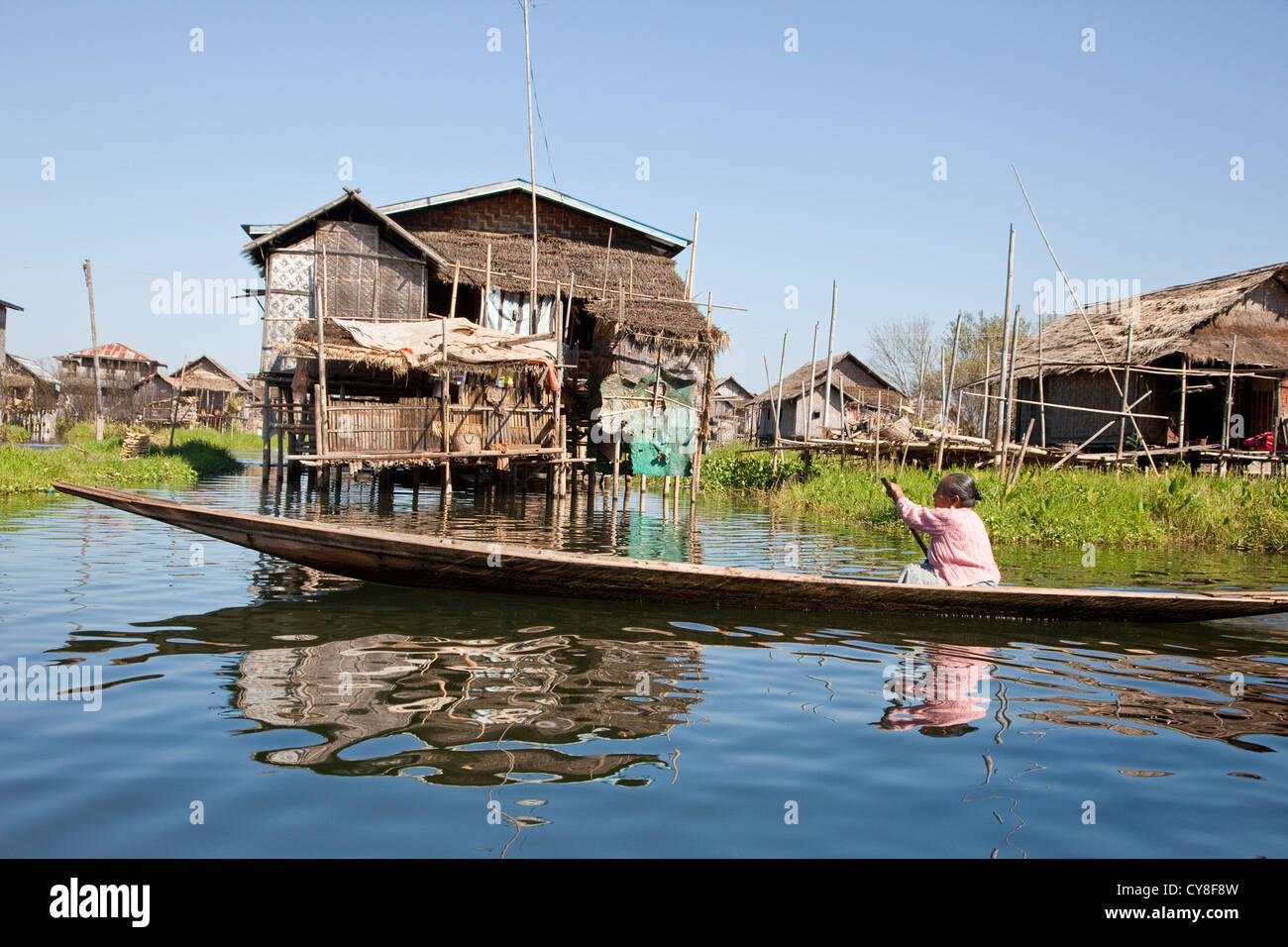 Le Myanmar, Birmanie. Scène de village, femme, bateau à rames, maisons sur pilotis, au Lac Inle, l'État Shan. Banque D'Images