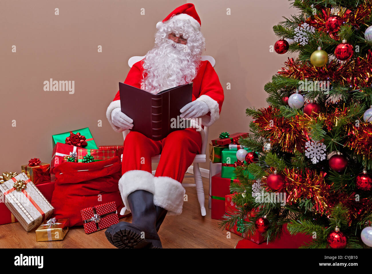 Santa Claus était assis dans une chaise à bascule la lecture de la 'liste' Gentil ou méchant entouré de gift wrapped presents. Banque D'Images