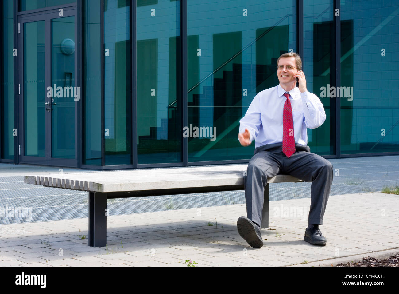 Businessman assis sur un banc en face d'un immeuble de bureaux et talking on mobile phone Banque D'Images