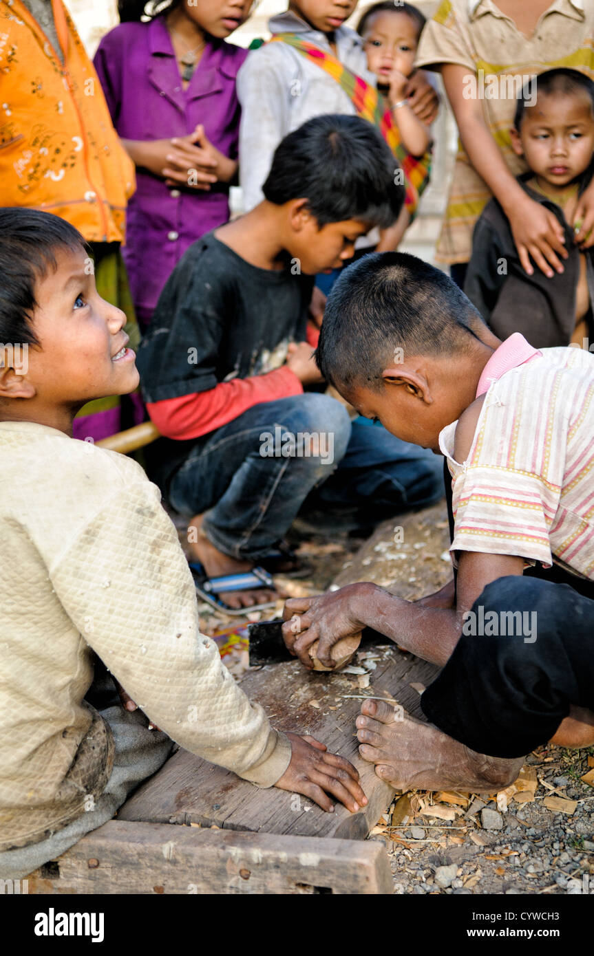 LUANG NAMTHA, Laos — Un jeune garçon dans un village de la province de Luang Namtha, au nord du Laos, sculpte soigneusement une toupie en bois pour le jeu traditionnel du tujlub. Il incorpore ingénieusement une pointe en métal d'une colonne vertébrale de parapluie pour créer un point durable pour ce jouet fabriqué à la main utilisé dans un jeu de haut de gamme compétitif lancé à la corde populaire parmi les enfants de la région. Banque D'Images