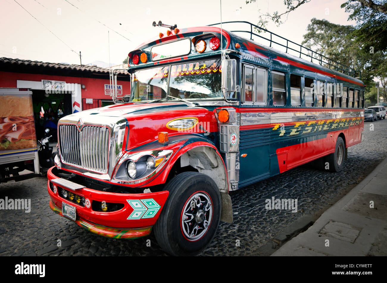 Chicken bus Antigua Guatemala Cobblestone Street | ANTIGUA, Guatemala — Un autobus scolaire américain à la retraite, repeint en chrome et cobalt avec le nom « Esmeralda » sur son flanc, est stationné dans une rue pavée. Les propriétaires de bus guatémaltèques achètent des unités Bluebird et International désaffectées et les personnalisent avec des noms, des chromes et des schémas de peinture folk-art, transformant chacun en une publicité roulante pour son itinéraire. Banque D'Images