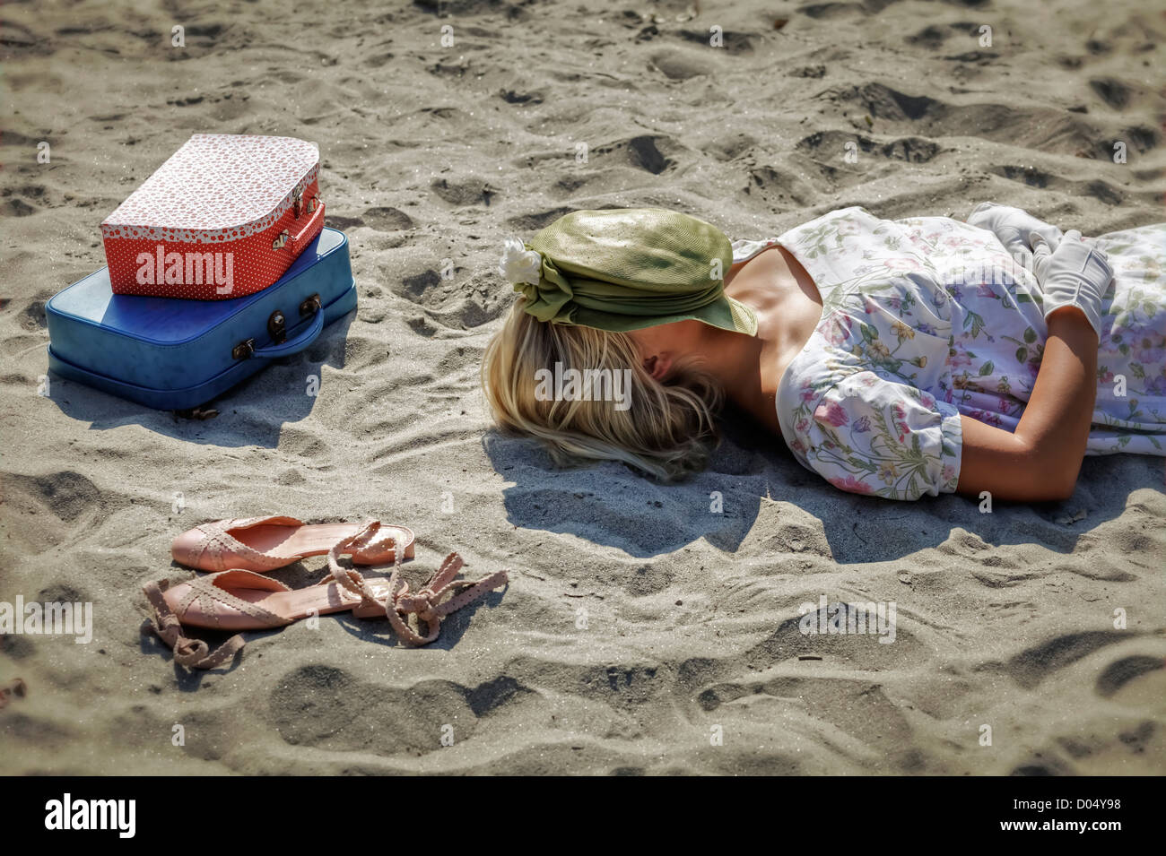 Une femme dans une robe florale est allongé sur une plage à côté de ses chaussures et deux valises vintage Banque D'Images