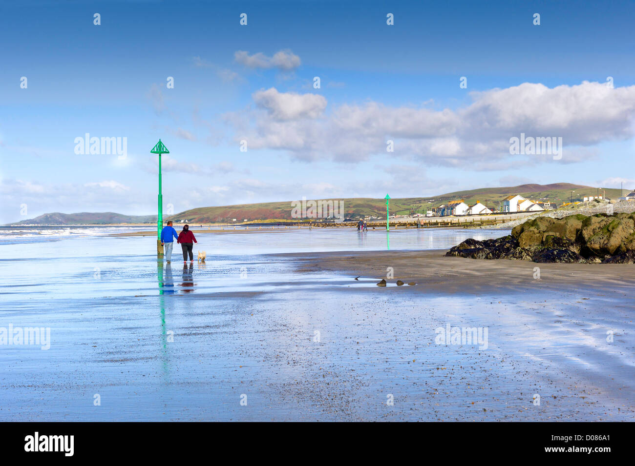 Mâles et femelles adultes avec un chien à pied sur une plage humide de la marée descendante, les vagues, les collines au loin, maisons en bord de mer Banque D'Images