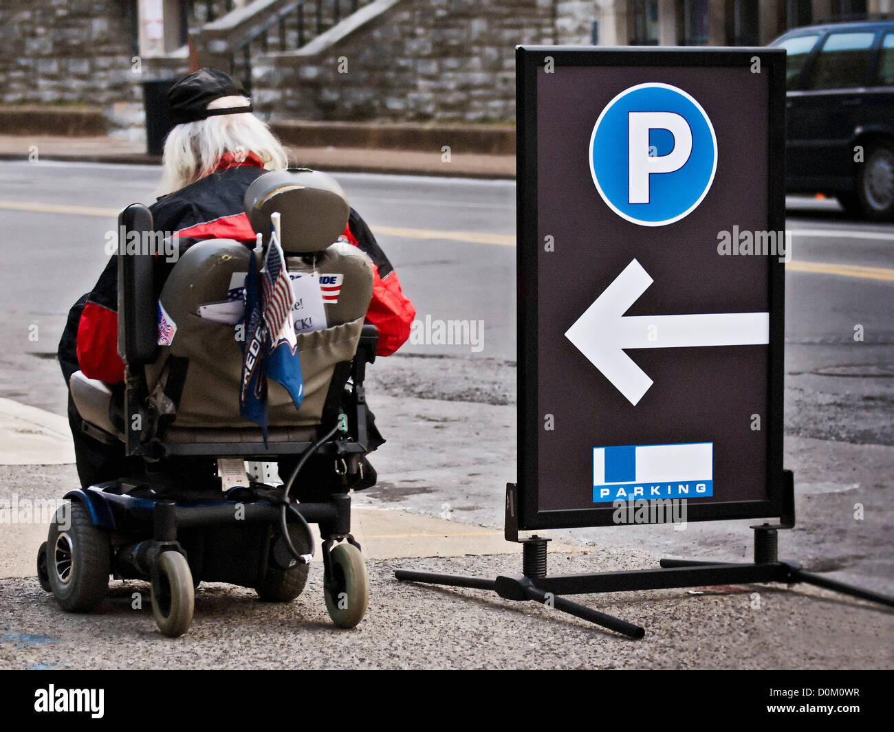 Un homme âgé dans un fauteuil roulant motorisé rouleaux passé un stationnement signe. Banque D'Images
