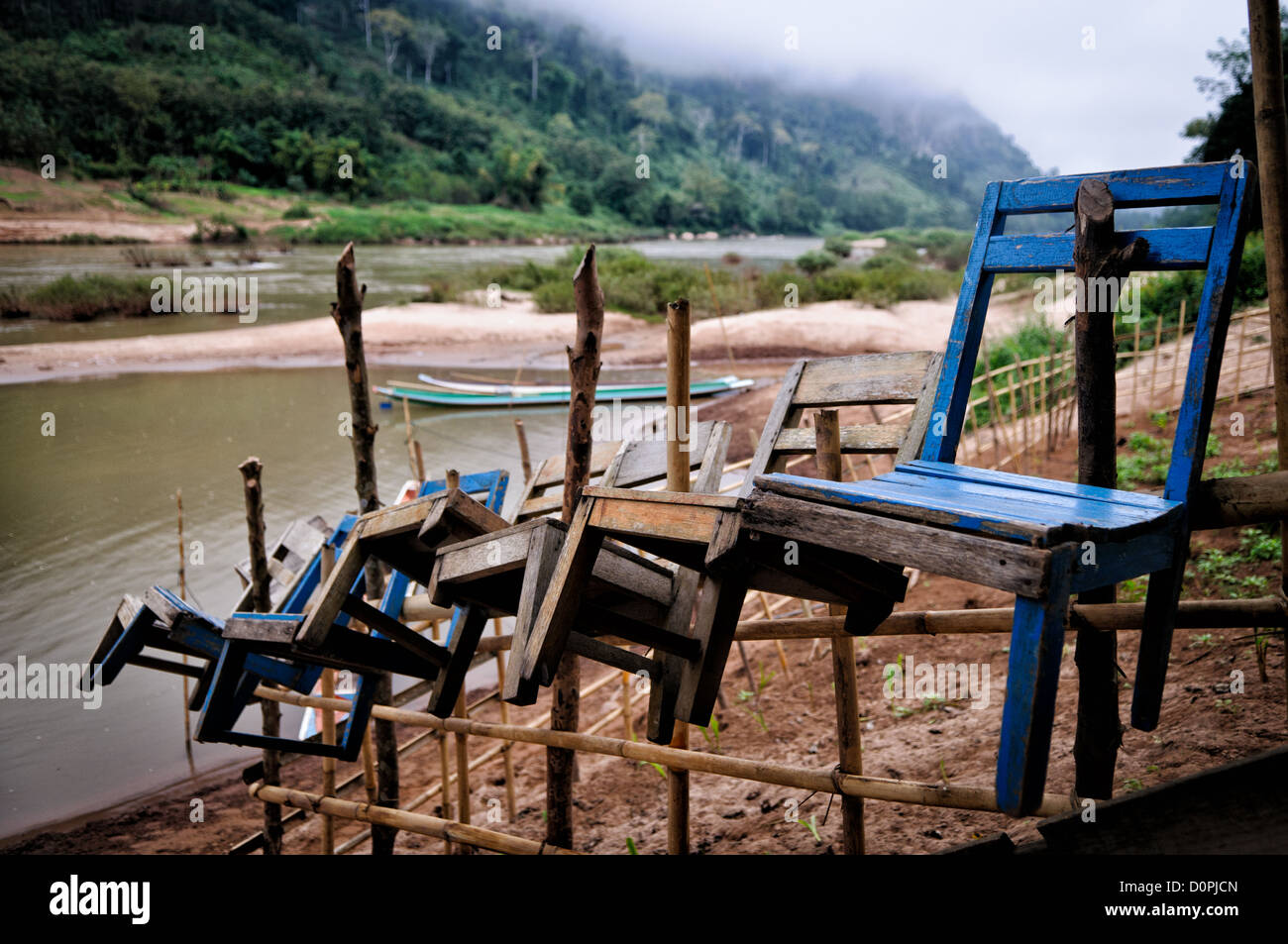 NONG KHIAW, Laos - Un groupe de chaises en bois sont suspendus sur des piquets en bois entourant un petit jardin sur les rives de la rivière Nam Ou (UO) de Nong Khiaw dans le nord de Laos. Dans l'arrière-plan, brumes matinales toujours impossibles à certains de le terrain accidenté de la campagne environnante. Banque D'Images