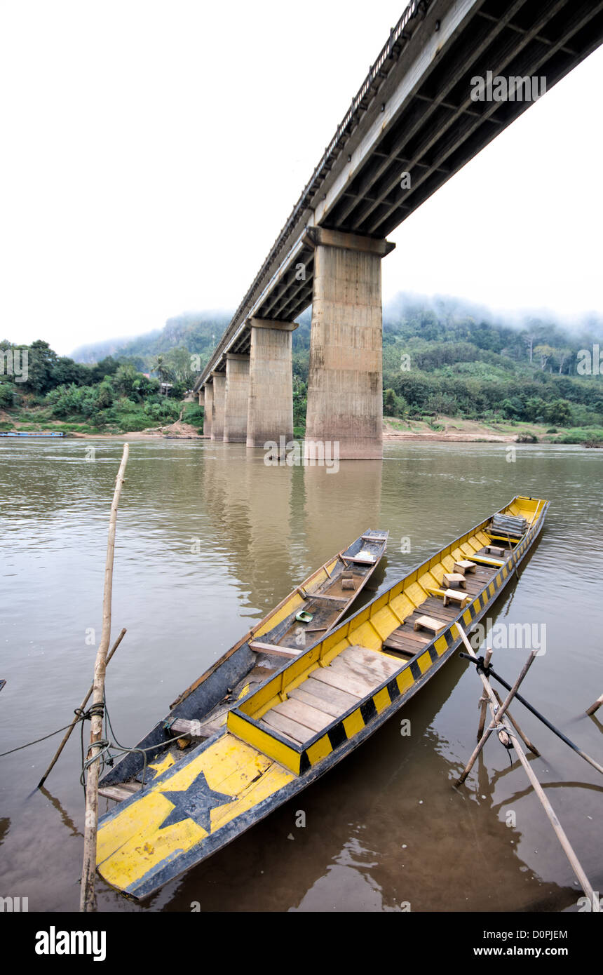 NONG KHIAW, Laos - open-top en bois bateaux attaché à un poteau sur les rives de la rivière Nam Ou (or) directement sous le haut pont en béton, enjambant la rivière de Nong Khiaw dans le nord du Laos. Banque D'Images