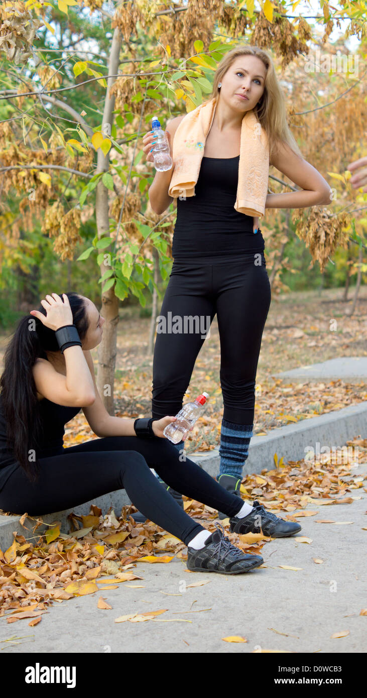 Deux athlètes féminines et de l'eau potable au repos assis sur le côté d'un chemin rural dans leurs vêtements de la réhydratation après une lourde Banque D'Images