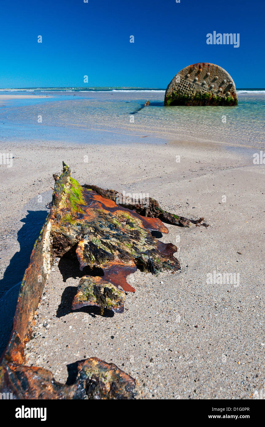 Métal rouillé, à demi-enterré dans le sable sur une plage, avec un réservoir de rouille dans l'eau derrière elle ; le ciel bleu et le soleil. Banque D'Images