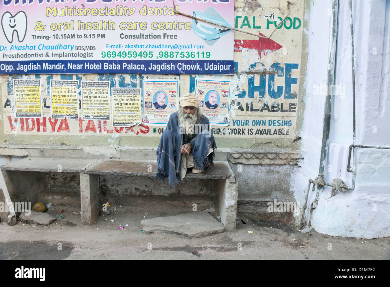 Un long longs cheveux Indiens barbu mendiant est assise en béton de l'AIIO banc en face des affiches publicitaires à Udaipur Rajasthan Inde Banque D'Images