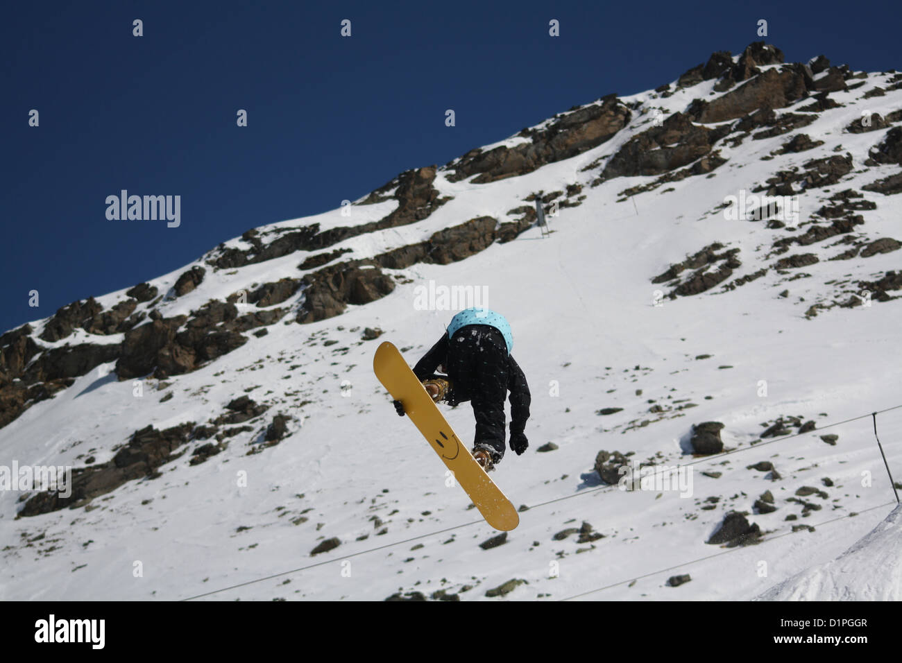 Male snowboarder dans l'air contre un décor de montagnes. Le snowboard est jaune avec un smiley face visible sur la face inférieure. Banque D'Images