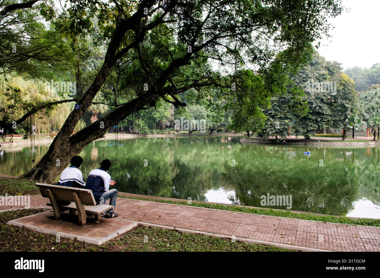 Hanoi Botanical Gardens Lake View Hanoi Vietnam // HANOI, Vietnam — deux jeunes hommes sont assis sur un banc donnant sur un petit lac dans les jardins botaniques de Hanoi, connu localement sous le nom de Vuon Bach Thao. Les jardins botaniques servent d'espace vert dans la capitale du Vietnam, offrant aux résidents et aux visiteurs des terrains paysagers et des plans d'eau. Situés dans le quartier de Ba Dinh, les jardins ont été créés pendant la période coloniale française et restent une zone de loisirs populaire. L'établissement dispose de diverses collections de plantes et de sentiers pédestres dans son parc. Les jardins botaniques de Hanoi continuent Banque D'Images
