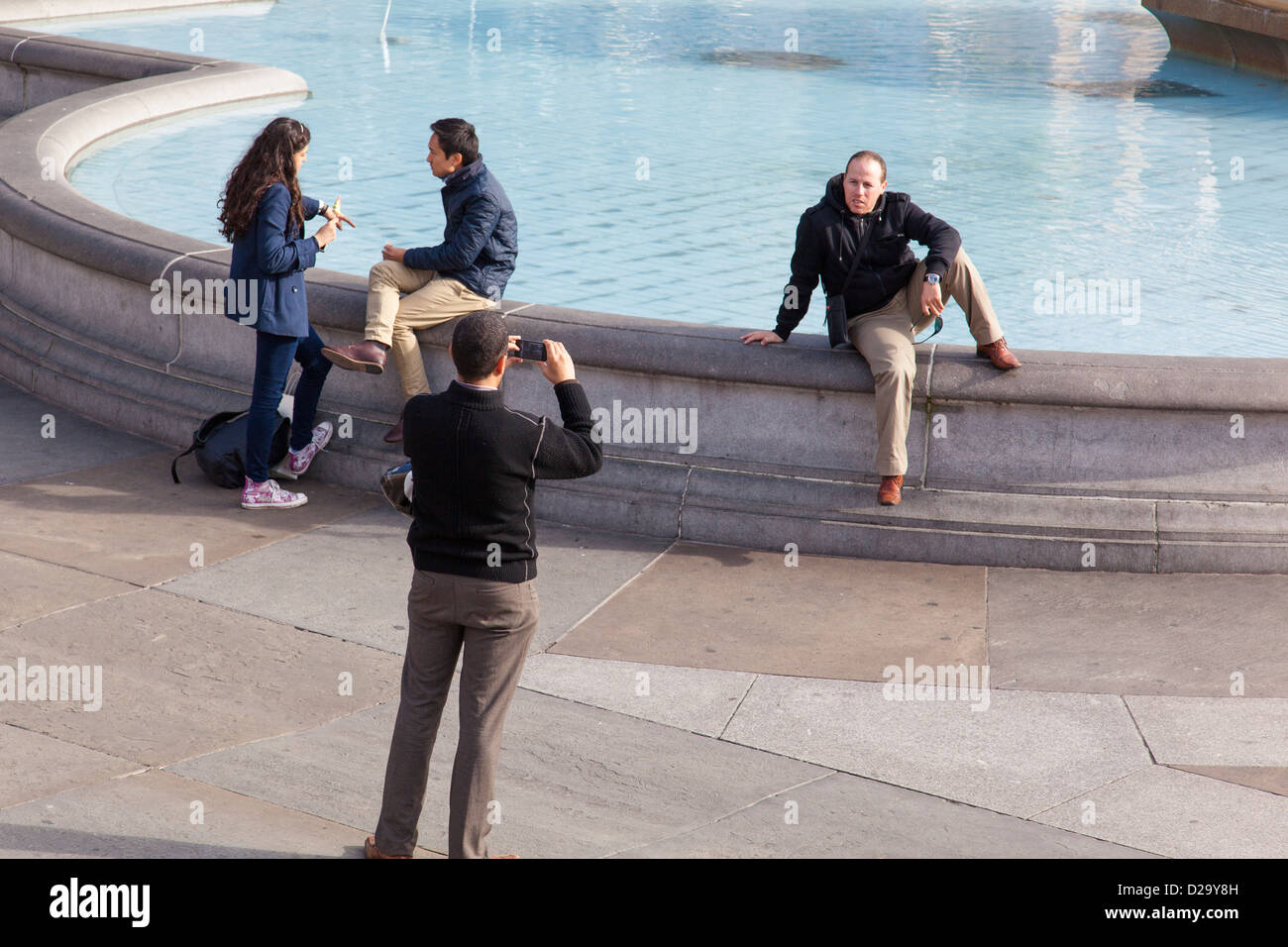 Tourist utilise son téléphone portable pour prendre une photo d'un homme assis sur le bord de la fontaine à Trafalgar Square, Londres. Banque D'Images