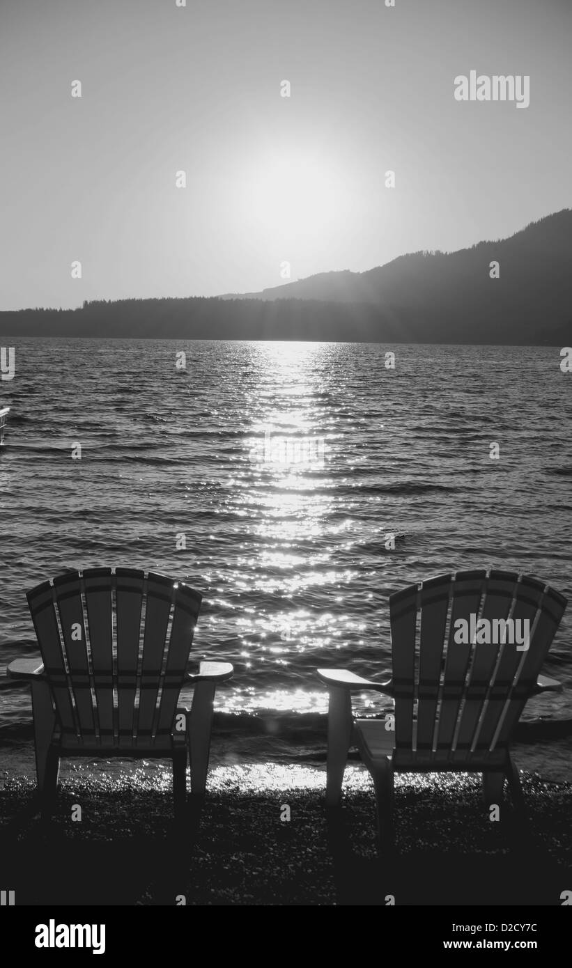 Image en échelle de gris de 2 chaises Adirondack sur une plage de galets avec le coucher de soleil au loin. Banque D'Images