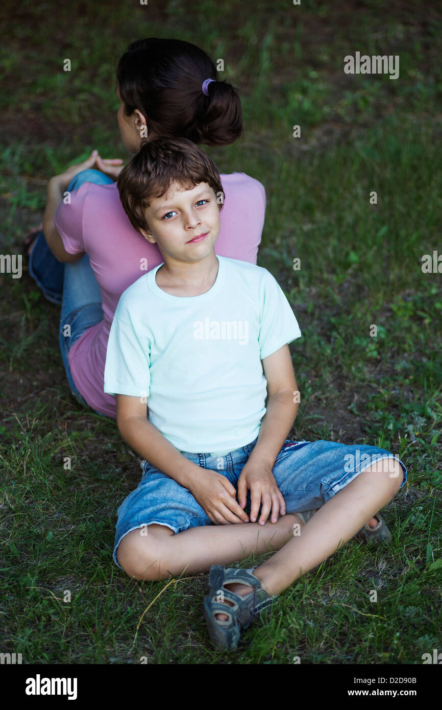 Un garçon assis en tailleur sur l'herbe, le dos appuyé contre le dos de sa maman Banque D'Images