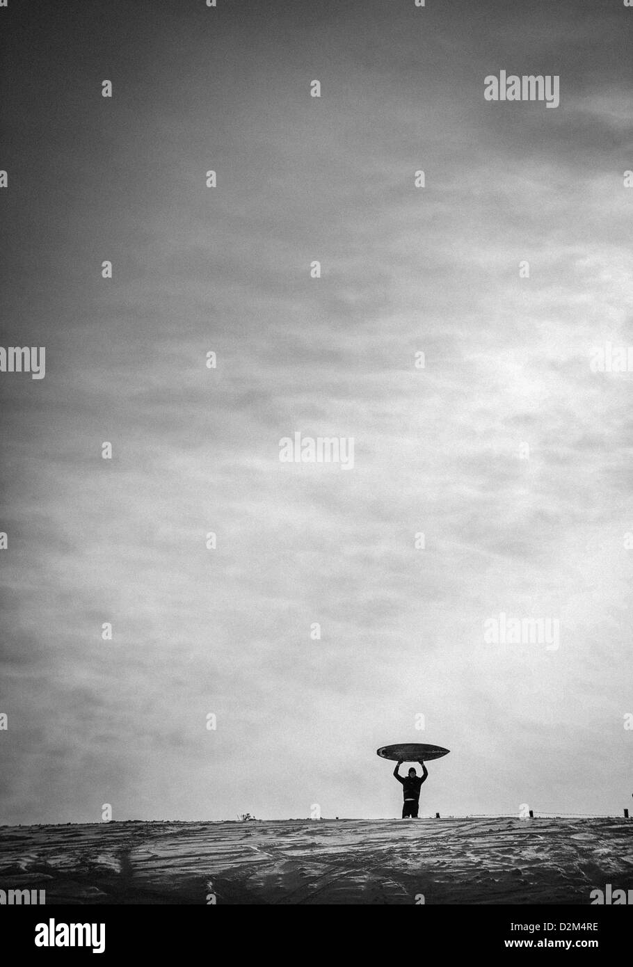 Un homme est titulaire d'une planche de surf au-dessus de sa tête en haut d'une colline couverte de neige près de Roseberry Topping dans Yorkshire, Angleterre. Banque D'Images
