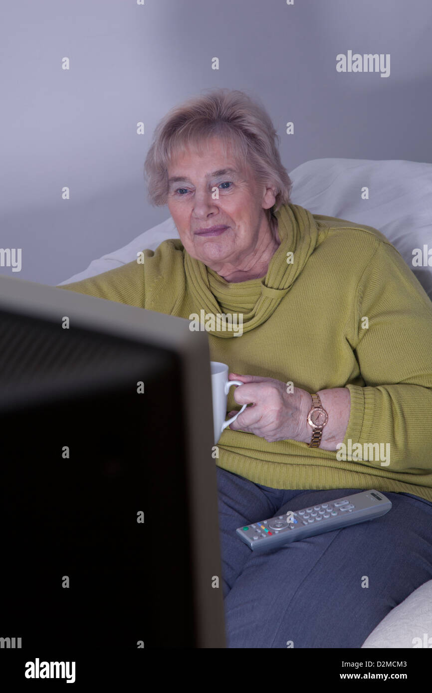 Mature femme assise tenant l'un mug de thé ou de café, regarder la télévision. Banque D'Images