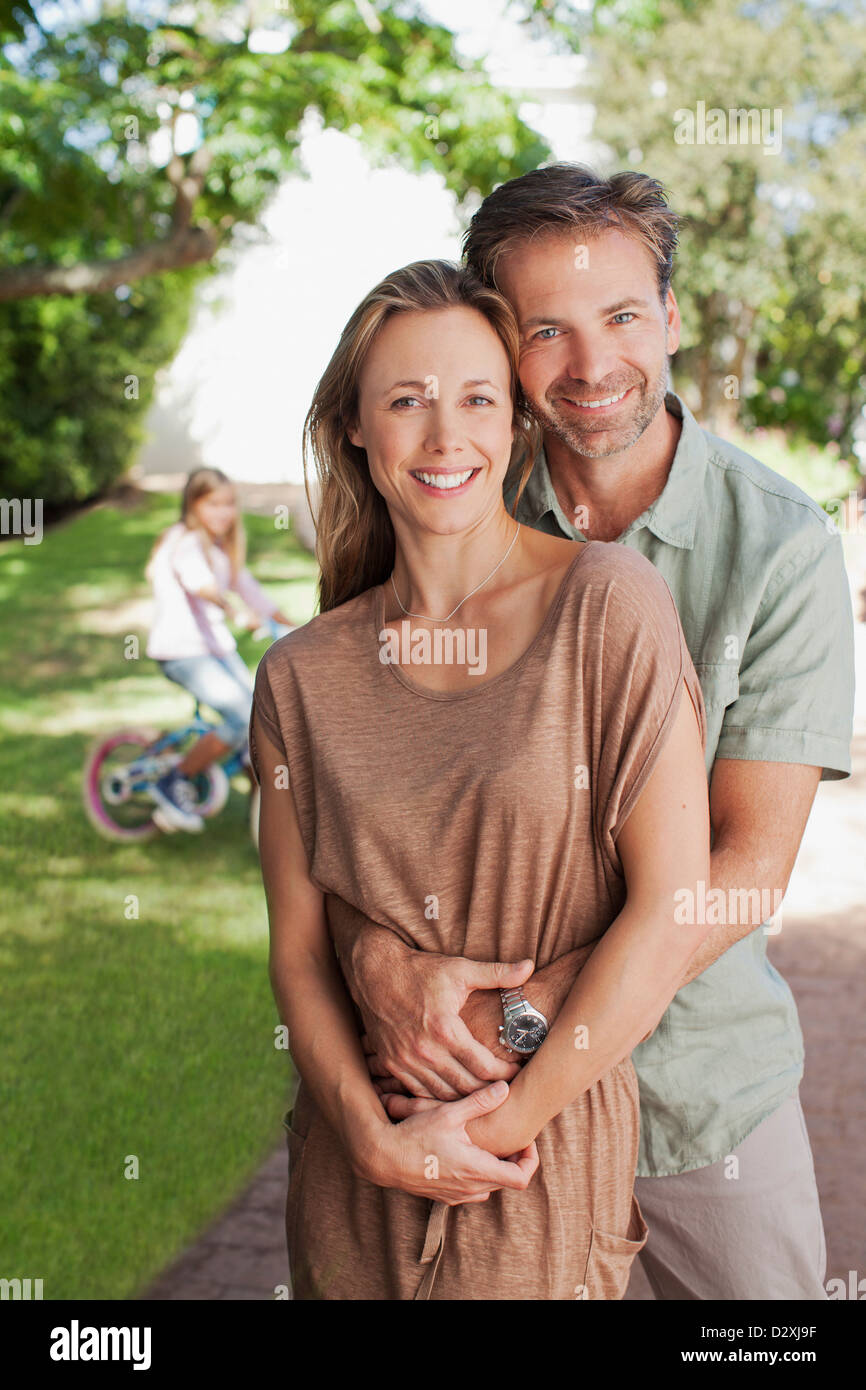 Portrait of smiling couple hugging in allée avec sa fille en bicyclette en arrière-plan Banque D'Images