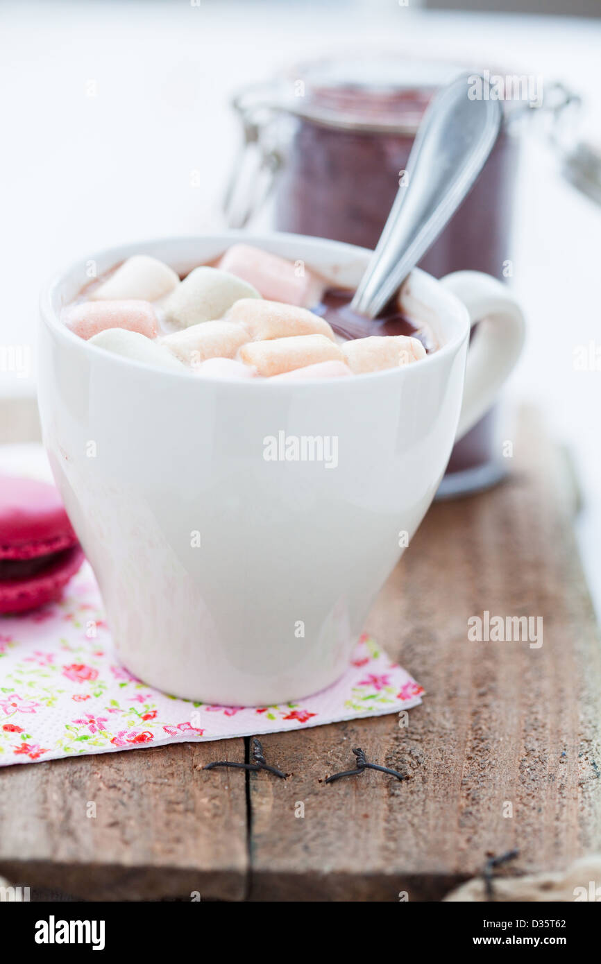 Libre de tasse de chocolat chaud avec guimauves colorés en appui sur table en bois avec un bocal en verre de poudre de cacao en arrière-plan Banque D'Images