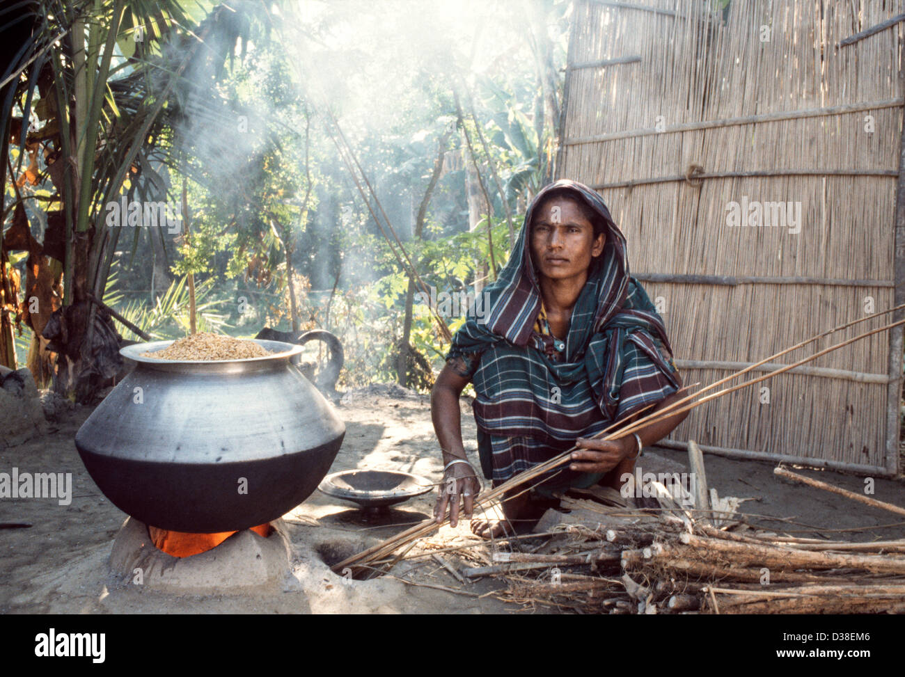 Village femme qui s'accroupira, nourrissant son poêle en argile ouvert avec du bois quand on fait du riz dans une casserole en métal à l'extérieur de sa maison. Près de Madaripur. Bangladesh Banque D'Images