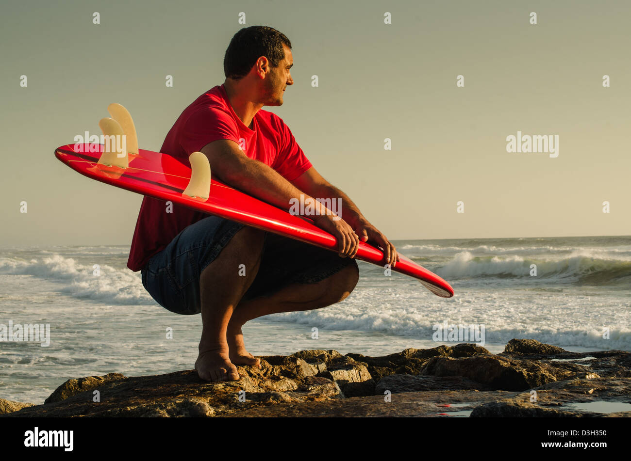 Un surfeur en regardant les vagues s'asseoir avec ses bras autour de son surf. Banque D'Images