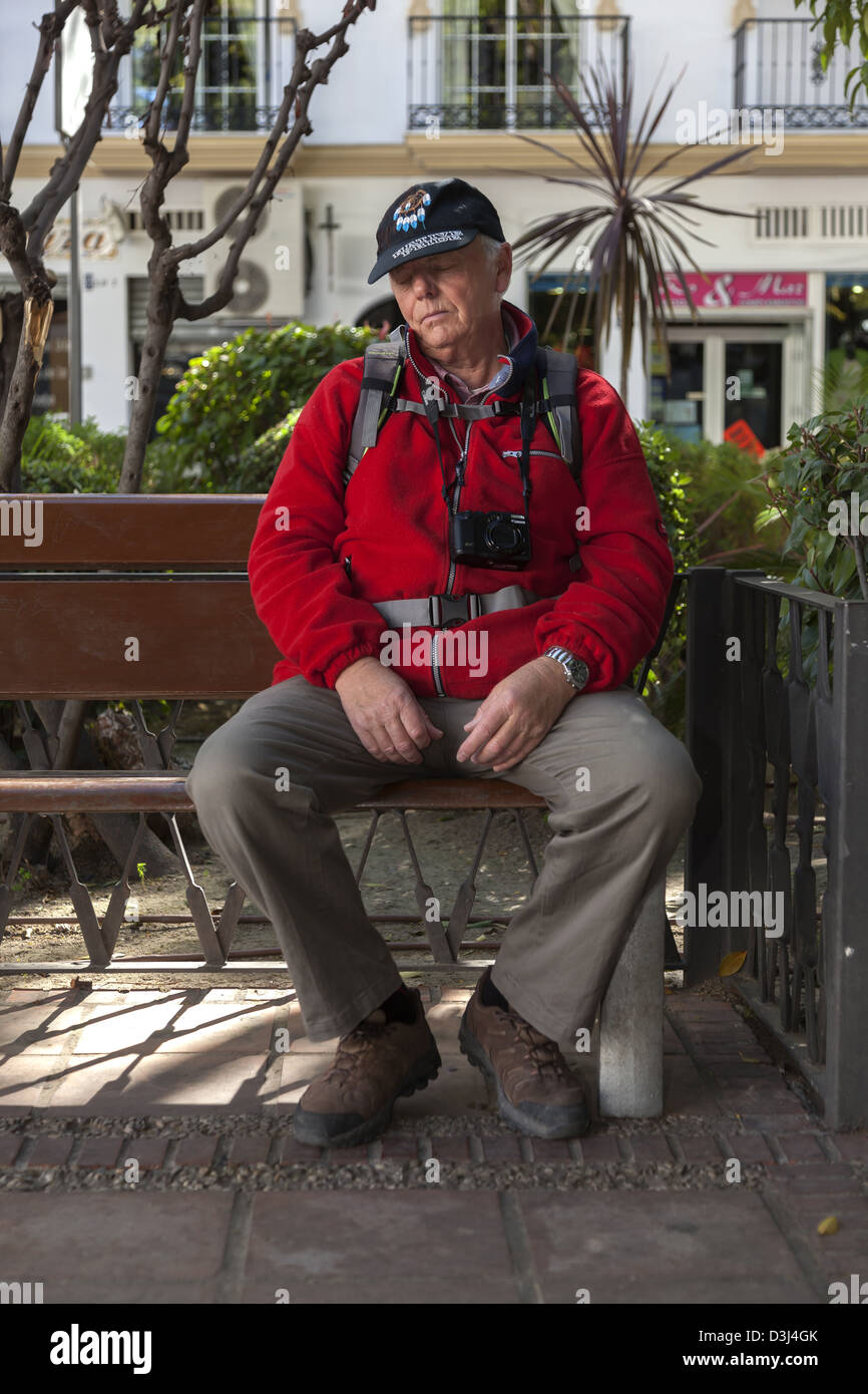 Senior male avec un appareil photo autour du cou et un sac à dos sonner pendant sur un banc de parc Banque D'Images